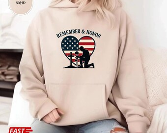 Puede incluir: Sudadera con capucha color arena con un gráfico en forma de corazón que presenta la bandera estadounidense y una silueta de soldado arrodillado. El texto "REMEMBER & HONOR" está encima. La sudadera tiene un bolsillo delantero.