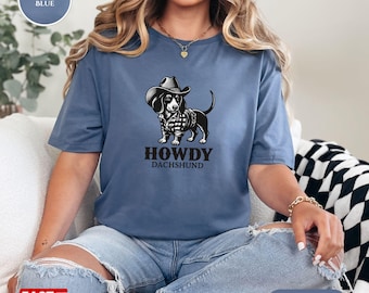 Könnte beinhalten: Indigoblaues T-Shirt mit einer Schwarz-Weiß-Illustration eines Dackels mit Cowboyhut und kariertem Hemd. Der Text "HOWDY DACHSHUND" steht unter dem Hund. Das Shirt ist ein Gildan 64000.