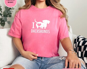 Könnte beinhalten: Ein pinkfarbenes T-Shirt mit dem weißen Text "I Dachshunds". Das Design zeigt ein Herz und eine Dackel-Silhouette. Das Shirt hat eine bequeme Farbe und eine lockere Passform.