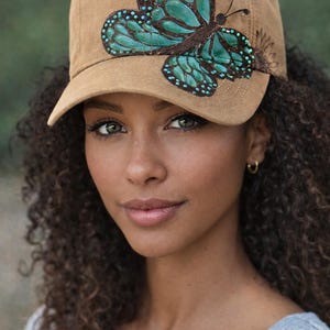 May include: Tan baseball cap featuring a large, teal and brown butterfly design on the front. The cap has a curved brim and a classic six-panel construction. A floral design is on the side of the cap.
