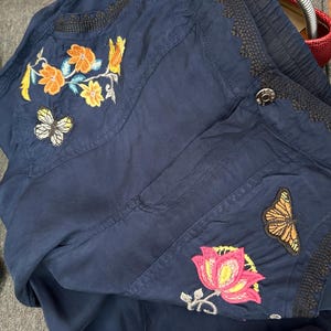May include: Navy blue jacket with embroidered floral and butterfly designs. The jacket features orange, yellow, pink, and white floral patterns, along with two butterfly patches. The jacket has a button closure and decorative trim.