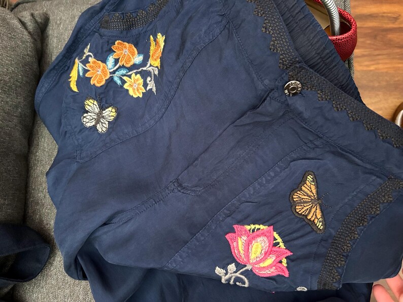 May include: Navy blue jacket with embroidered floral and butterfly designs. The jacket features orange, yellow, pink, and white floral patterns, along with two butterfly patches. The jacket has a button closure and decorative trim.