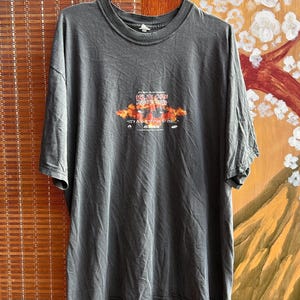 May include: Dark gray t-shirt with a graphic featuring the text "KLINGON FOREVER SURF" above an image of a can and the phrase "IT'S A GOOD DAY TO DIE..." in orange and red. The shirt has a crew neck and short sleeves.