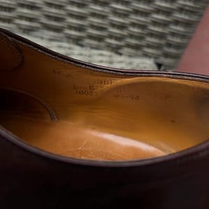 May include: Close-up of the interior of a burgundy leather shoe. The insole is a light brown color with visible markings, including size and style information. The shoe's stitching and construction are visible.
