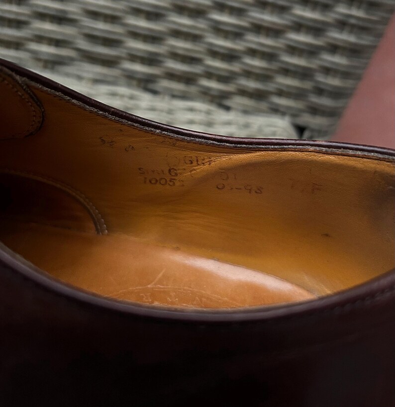 May include: Close-up of the interior of a burgundy leather shoe. The insole is a light brown color with visible markings, including size and style information. The shoe's stitching and construction are visible.