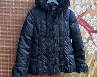 May include: Black puffer jacket with a faux fur-trimmed hood, cuffs, and front panel. The jacket features decorative stitching down the front and on the sleeves. The jacket is hanging on a black hanger.