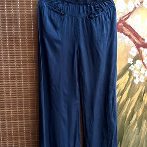 May include: Navy blue wide-leg trousers hanging on a black hanger. The trousers have a gathered waistband and a flowing, silky texture. The fabric drapes smoothly, suggesting a lightweight material. The trousers are displayed against a backdrop of a bamboo blind and a painted wall.