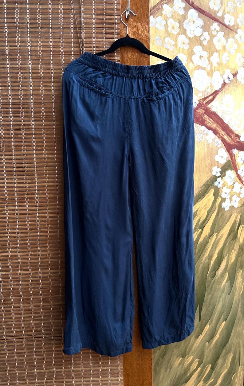 May include: Navy blue wide-leg trousers hanging on a black hanger. The trousers have a gathered waistband and a flowing, silky texture. The fabric drapes smoothly, suggesting a lightweight material. The trousers are displayed against a backdrop of a bamboo blind and a painted wall.