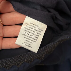 May include: A close-up of a navy blue garment tag with the text: "SELF: 60% CUPRA 40% RAYON, TRIMS: 100% POLYESTER, EXCLUSIVE OF DECORATION, MACHINE WASH COLD, ONLY NON-CHLORINE, BLEACH WHEN NEEDED, TUMBLE DRY LOW, WARM IRON, RN#149414, MADE IN CHINA."
