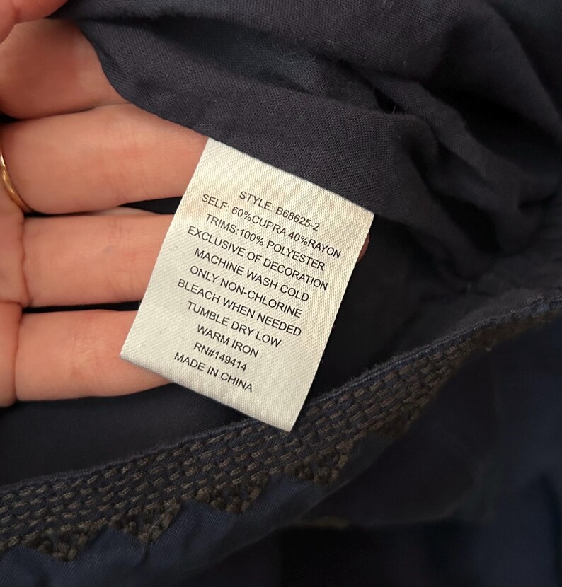 May include: A close-up of a navy blue garment tag with the text: "SELF: 60% CUPRA 40% RAYON, TRIMS: 100% POLYESTER, EXCLUSIVE OF DECORATION, MACHINE WASH COLD, ONLY NON-CHLORINE, BLEACH WHEN NEEDED, TUMBLE DRY LOW, WARM IRON, RN#149414, MADE IN CHINA."
