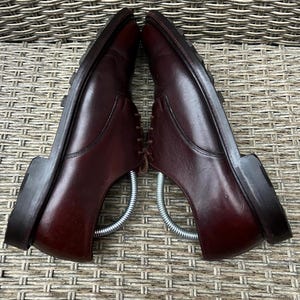 May include: Pair of burgundy leather dress shoes with laces and a rubber sole. The shoes are displayed with metal shoe trees inside, showcasing their shape and structure. The shoes are set against a textured background.