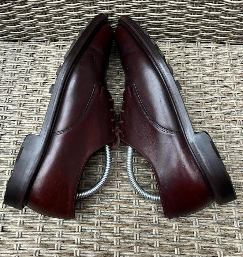 May include: Pair of burgundy leather dress shoes with laces and a rubber sole. The shoes are displayed with metal shoe trees inside, showcasing their shape and structure. The shoes are set against a textured background.