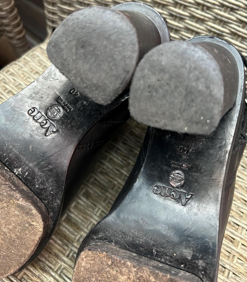 May include: Close-up of the soles of two black leather boots. The heels are worn and show signs of use. The brand name "Acne" is visible on the sole, along with the size 40.