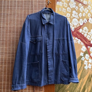May include: A dark blue denim chore jacket featuring a classic collar, a button-front closure, and four front pockets. The jacket has long sleeves with buttoned cuffs and a structured fit, showcasing a durable fabric texture and visible stitching details.