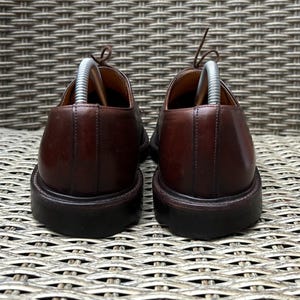 May include: A pair of brown leather dress shoes with laces and shoe trees inserted. The shoes have a rounded toe and a dark brown sole. The background is a woven wicker pattern.