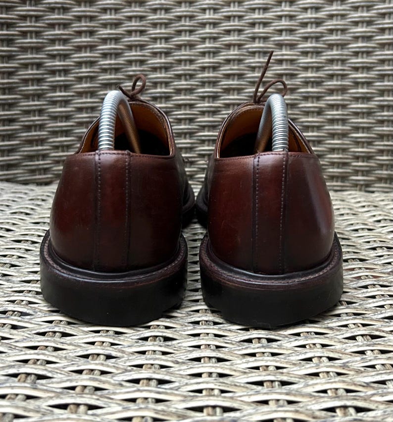May include: A pair of brown leather dress shoes with laces and shoe trees inserted. The shoes have a rounded toe and a dark brown sole. The background is a woven wicker pattern.