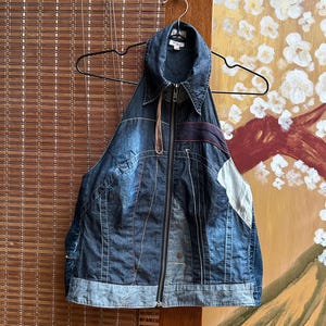 Peut inclure: Veste sans manches en jean avec fermeture éclair et col. La veste présente un design patchwork avec différentes nuances de denim bleu et des coutures contrastées. Elle est suspendue à un cintre noir devant un store en bambou.