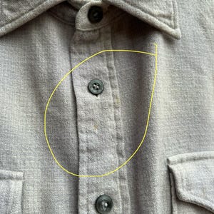 以下が含まれることがあります： 2つの胸ポケットが付いた、明るい灰色のボタンダウンシャツのクローズアップ。 シャツには襟があり、黒いボタンでボタン留めされています。 生地は、テクスチャード加工された織り素材のようです。