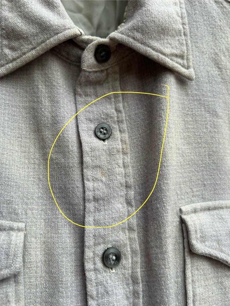 以下が含まれることがあります： 2つの胸ポケットが付いた、明るい灰色のボタンダウンシャツのクローズアップ。 シャツには襟があり、黒いボタンでボタン留めされています。 生地は、テクスチャード加工された織り素材のようです。