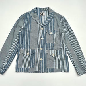 Puede incluir: Chaqueta a rayas azules y blancas con cuello chal y cuatro bolsillos delanteros. La chaqueta tiene cierre de botones y una pequeña etiqueta cerca del cuello. Las mangas son largas y la tela parece ser una mezcla ligera de algodón o lino.