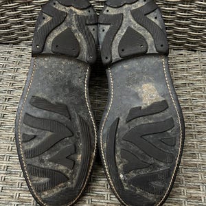 May include: A close-up shot of the soles of two worn leather boots. The soles are black with a textured, patterned design and show signs of wear. The stitching is visible along the edges of the soles.