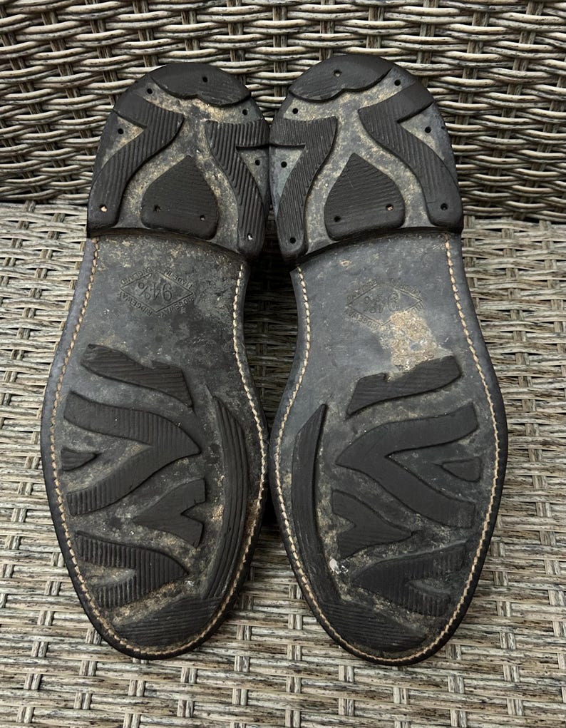 May include: A close-up shot of the soles of two worn leather boots. The soles are black with a textured, patterned design and show signs of wear. The stitching is visible along the edges of the soles.