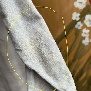以下が含まれることがあります： 明るい灰色の長袖シャツの袖のクローズアップ。生地は質感のある外観です。袖にシミがあります。背景は白い花のある絵画のぼやけた画像です。
