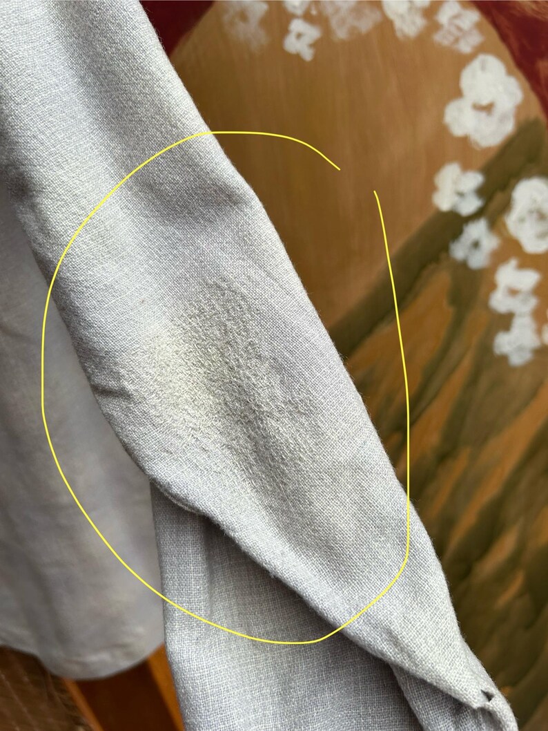 以下が含まれることがあります： 明るい灰色の長袖シャツの袖のクローズアップ。生地は質感のある外観です。袖にシミがあります。背景は白い花のある絵画のぼやけた画像です。
