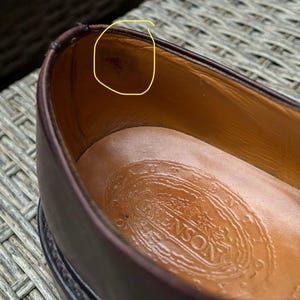 May include: Close-up of a brown leather shoe interior. The shoe's interior is tan, with a stamped logo on the insole. The shoe's exterior is a dark brown color. The shoe appears to be well-worn.