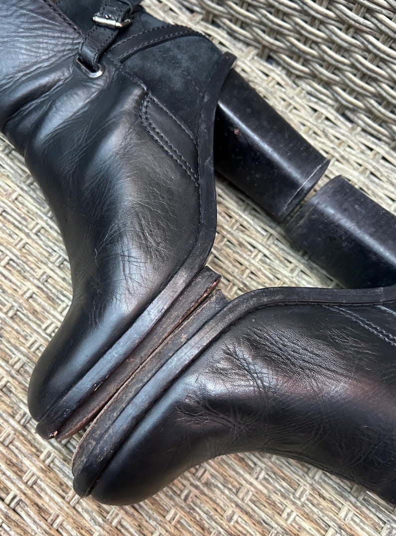 May include: Black leather boots with a high heel and a buckle detail. The boots have a worn appearance, with visible wear on the soles and heels. The heel is approximately 4 inches.