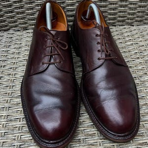 May include: A pair of dark burgundy leather dress shoes with a classic lace-up design. The shoes have a polished finish and are displayed with shoe trees inside. The soles are dark brown, and the shoes are set against a textured background.