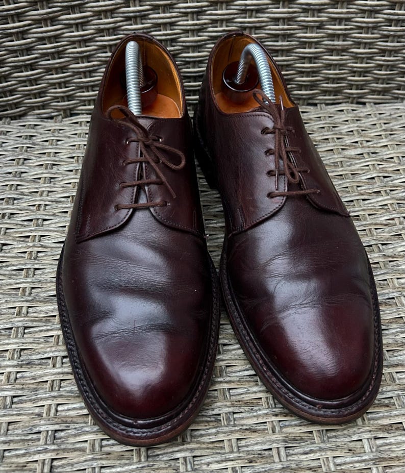 May include: A pair of dark burgundy leather dress shoes with a classic lace-up design. The shoes have a polished finish and are displayed with shoe trees inside. The soles are dark brown, and the shoes are set against a textured background.