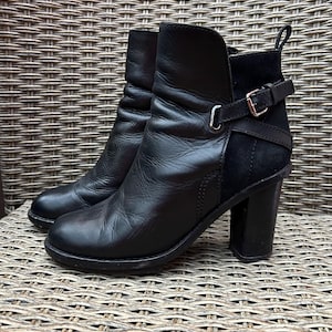 May include: A pair of black leather ankle boots with a block heel. The boots have a rounded toe, a short shaft, and a strap with a silver buckle. The boots are sitting on a woven surface.
