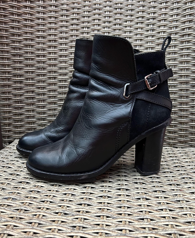 May include: A pair of black leather ankle boots with a block heel. The boots have a rounded toe, a short shaft, and a strap with a silver buckle. The boots are sitting on a woven surface.