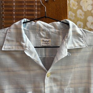 May include: A light blue and white plaid button-down shirt with a pointed collar and a single visible button. The interior neck label reads Pennleigh WASH 'N WEAR 15 1/2 33. The shirt features a horizontal and vertical striped pattern throughout.