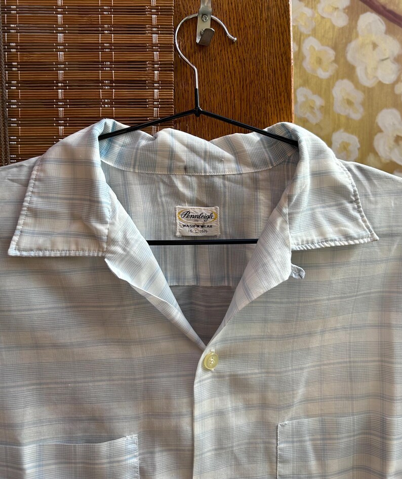 May include: A light blue and white plaid button-down shirt with a pointed collar and a single visible button. The interior neck label reads Pennleigh WASH 'N WEAR 15 1/2 33. The shirt features a horizontal and vertical striped pattern throughout.