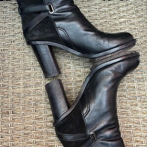 May include: Black leather ankle boots with a block heel. The boots have a strap detail with a buckle and a suede panel. The boots are displayed on a woven surface.