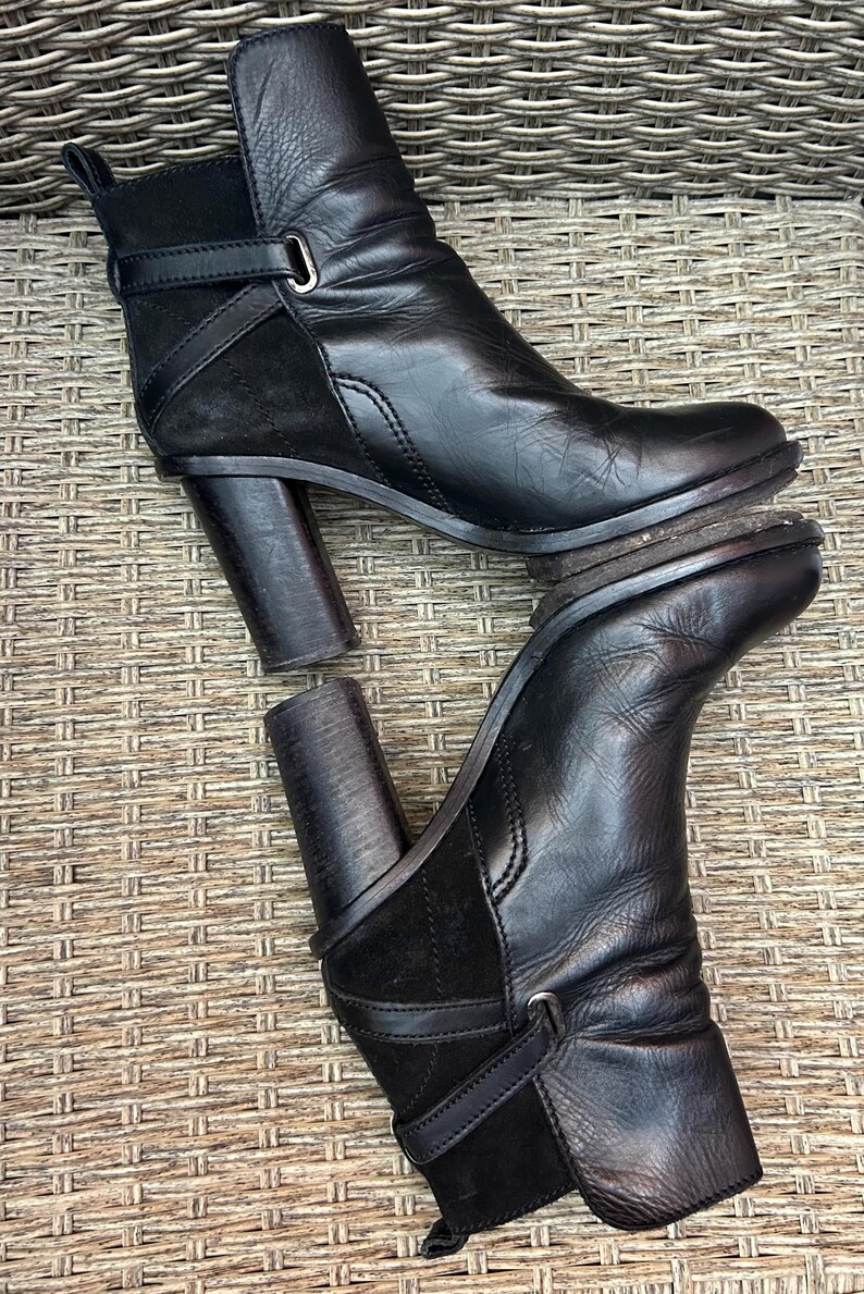 May include: Black leather ankle boots with a block heel. The boots have a strap detail with a buckle and a suede panel. The boots are displayed on a woven surface.