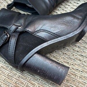 May include: Close-up of a black leather boot with a block heel. The boot features a buckle detail and a suede panel. The leather appears worn, and the heel is approximately 3 inches high.