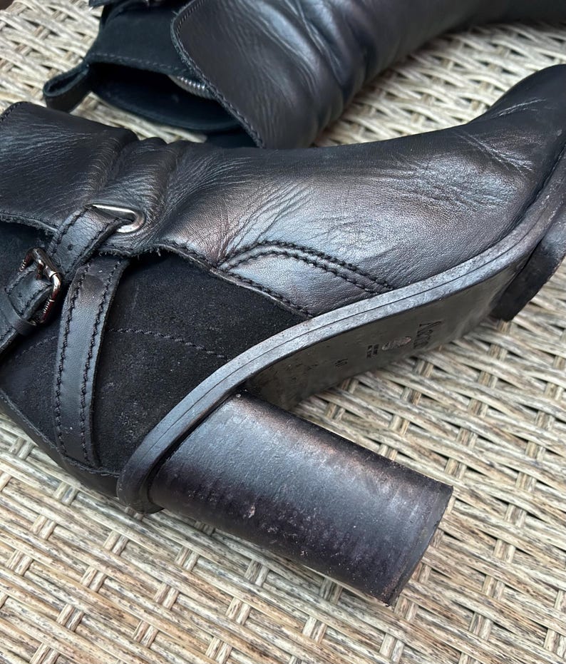 May include: Close-up of a black leather boot with a block heel. The boot features a buckle detail and a suede panel. The leather appears worn, and the heel is approximately 3 inches high.