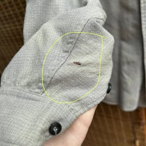以下が含まれることがあります： 明るい灰色の長袖シャツの袖のクローズアップ。 袖口には黒いボタンが付いています。 袖口の近くには小さな茶色のシミがあります。