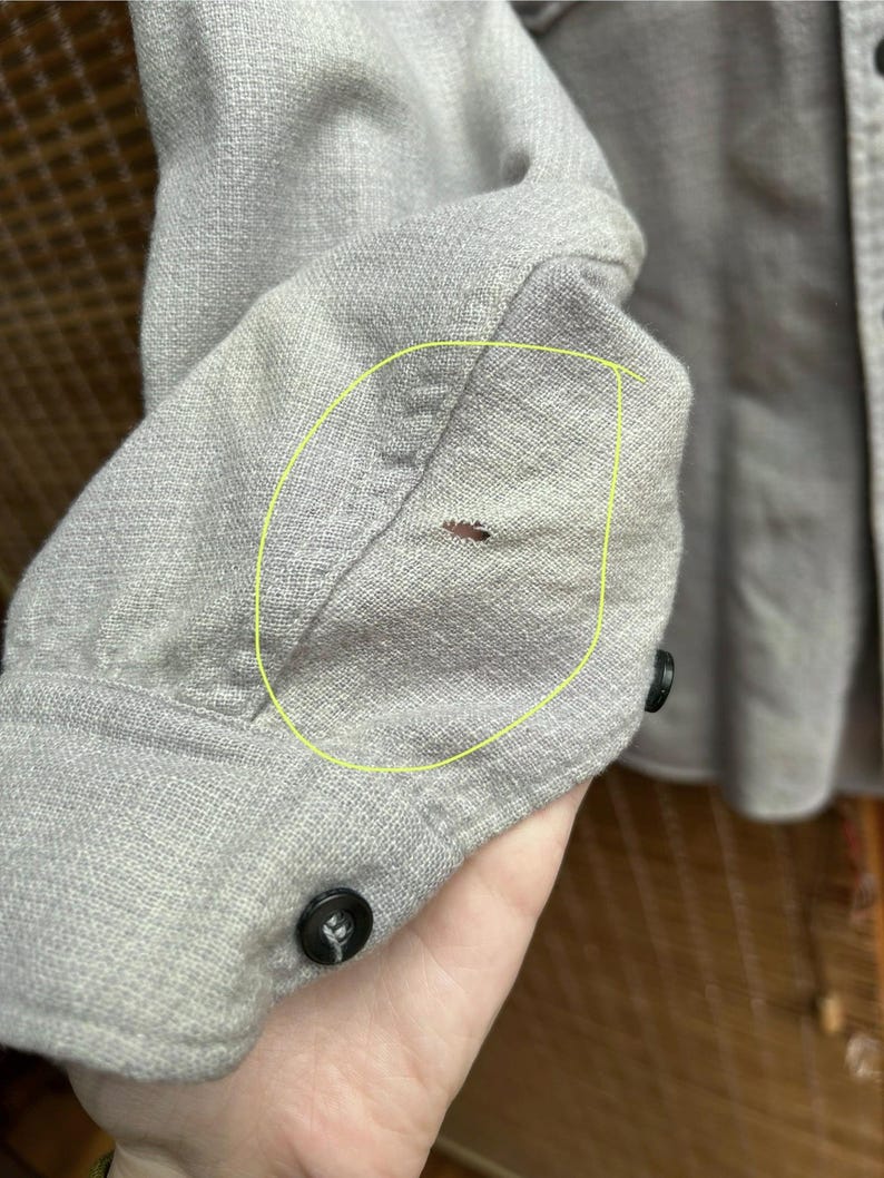 以下が含まれることがあります： 明るい灰色の長袖シャツの袖のクローズアップ。 袖口には黒いボタンが付いています。 袖口の近くには小さな茶色のシミがあります。