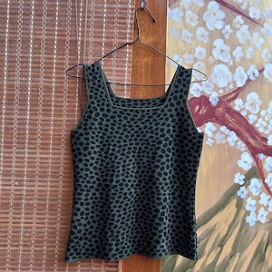 Pode incluir: Uma regata verde-oliva com um padrão de animal preto. A parte superior tem um decote quadrado e alças largas. Está pendurada em um cabide de metal contra um fundo de uma persiana de bambu e uma parede pintada.