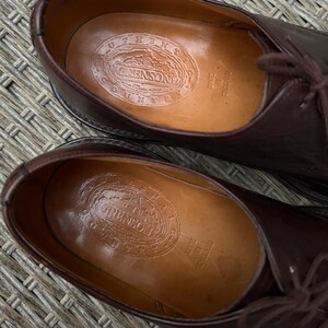 May include: Close-up of a pair of brown leather dress shoes. The interior of each shoe displays a stamped logo that reads "GRENSON COUNTY" and "MADE IN ENGLAND". The shoes have laces and a classic design.