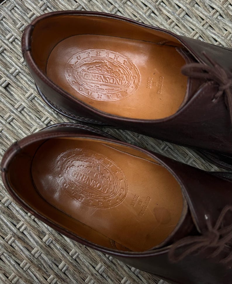 May include: Close-up of a pair of brown leather dress shoes. The interior of each shoe displays a stamped logo that reads "GRENSON COUNTY" and "MADE IN ENGLAND". The shoes have laces and a classic design.
