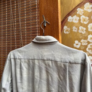 以下が含まれることがあります： 金属製のフックに掛けられた、明るい灰色のボタンダウンシャツ。 シャツには襟とテクスチャード加工の生地があります。 背景には、竹製のブラインド、木のドア、白い花のアクセントが付いた金色の絵画が含まれています。