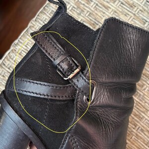 May include: Close-up of a black leather boot with a suede panel and a buckle detail. The boot features a small strap with a silver-colored buckle. The leather appears to be textured, and the suede panel is stitched.