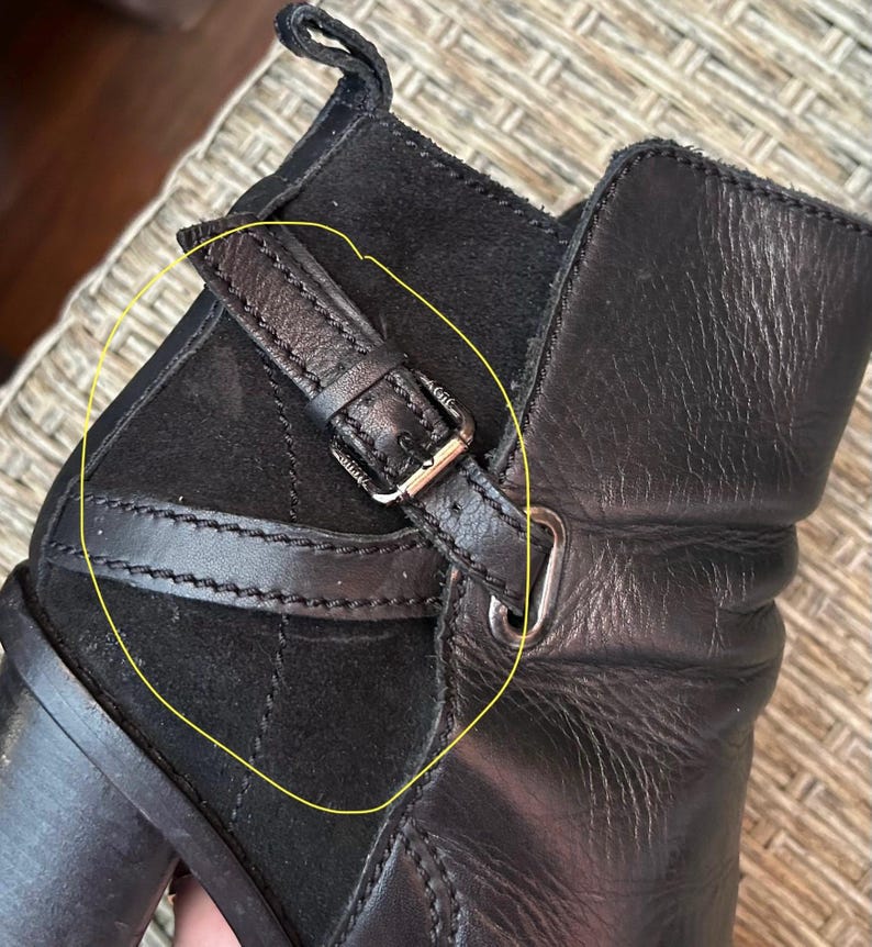 May include: Close-up of a black leather boot with a suede panel and a buckle detail. The boot features a small strap with a silver-colored buckle. The leather appears to be textured, and the suede panel is stitched.