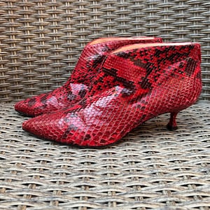 Puede incluir: Un par de botines con un vibrante patrón de piel de serpiente en rojo y negro. Estos botines presentan una punta afilada, un tacón bajo de tipo kitten y una silueta elegante que llega justo por encima del tobillo, con un acabado texturizado.