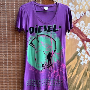 Könnte beinhalten: Lila Diesel T-Shirt mit Rundhalsausschnitt und kurzen Ärmeln. Das Shirt zeigt eine Grafik eines Frauenprofils in Schwarz und Grün, mit dem Wort "DIESEL" darüber. Zusätzlicher Text und florale Akzente sind ebenfalls vorhanden.